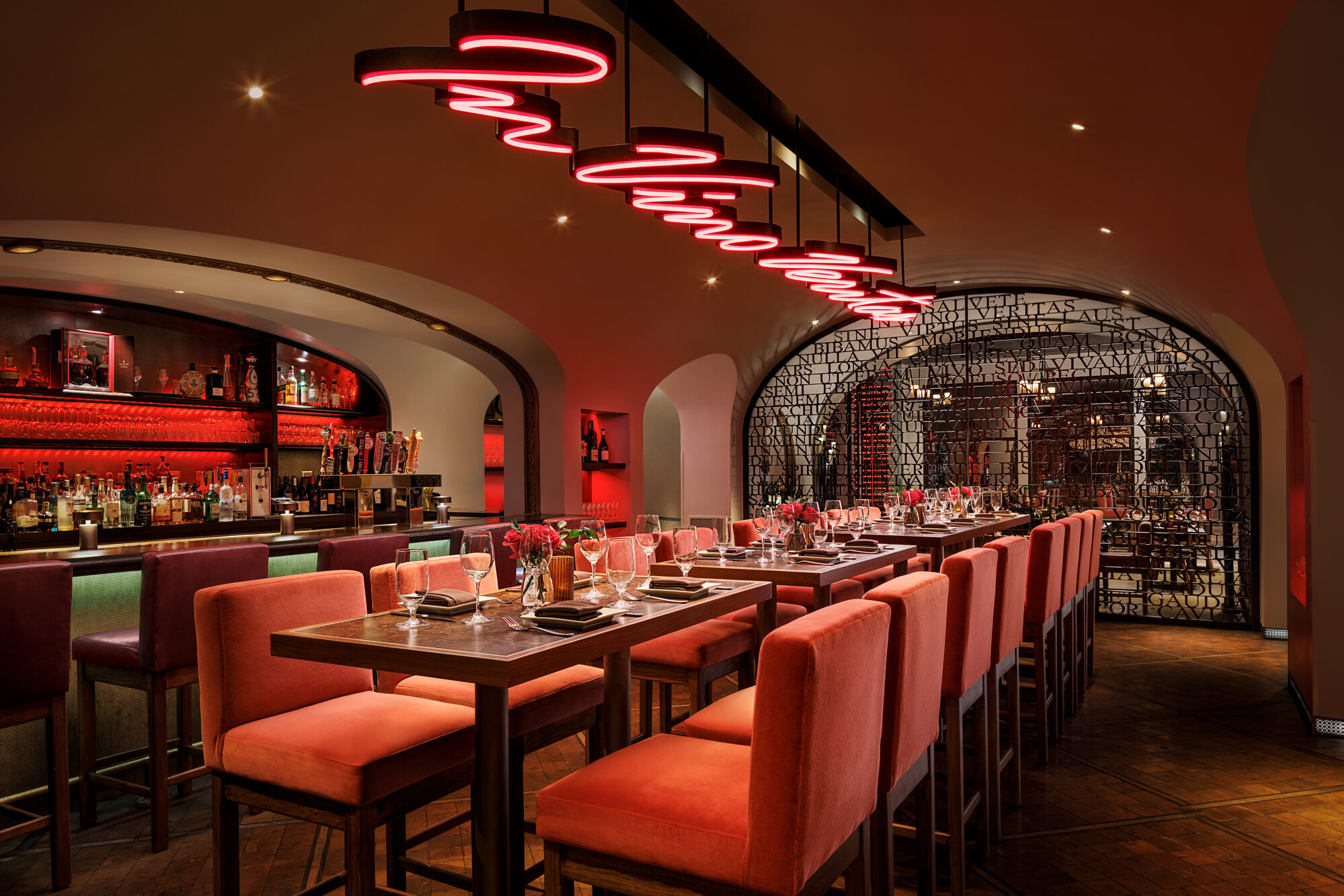 The Cellar's long communal table beneath coiled neon lighting, leading toward the arched wine gate with the 'In Vino Veritas' iron screen