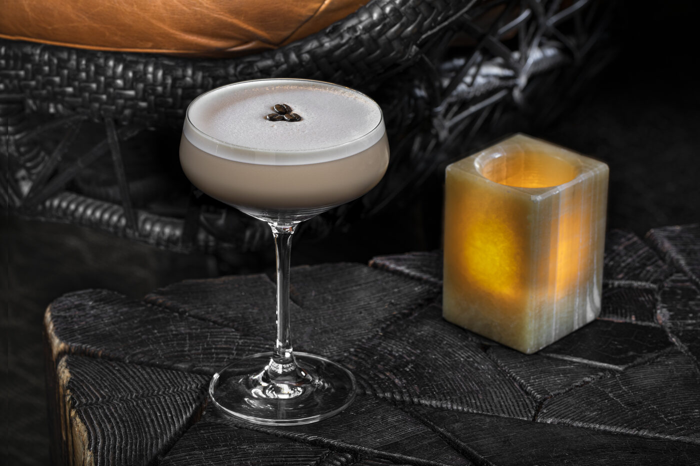 Espresso Martini in a coupe with silky crema and three coffee beans, beside a glowing onyx candle on a dark woven ottoman